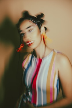 The image depicts a blurred portrait of a person. The individual has an artistic hairstyle with buns and wears a colorful, vertically striped sleeveless top. The background is a warm, muted tone. The blur effect creates a dynamic and abstract feeling.