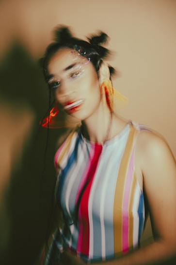 The image depicts a blurred portrait of a person. The individual has an artistic hairstyle with buns and wears a colorful, vertically striped sleeveless top. The background is a warm, muted tone. The blur effect creates a dynamic and abstract feeling.