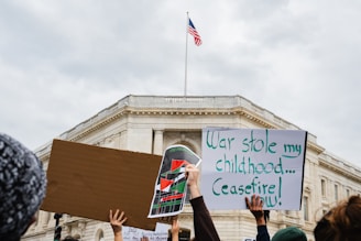 Several people are holding protest signs in front of a large government building. One sign is blank, another features a colorful design with flags, and the third reads 'War stole my childhood... Ceasefire now!' An American flag is flying atop the building.