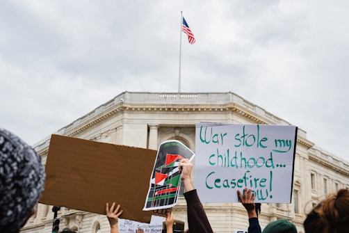 Several people are holding protest signs in front of a large government building. One sign is blank, another features a colorful design with flags, and the third reads 'War stole my childhood... Ceasefire now!' An American flag is flying atop the building.