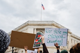 Several people are holding protest signs in front of a large government building. One sign is blank, another features a colorful design with flags, and the third reads 'War stole my childhood... Ceasefire now!' An American flag is flying atop the building.