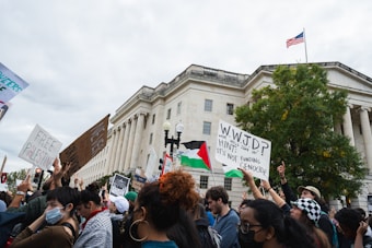 A group of protestors gather in front of a large government building holding various signs and flags. Signs contain messages supporting Palestine and opposing funding for genocide. The crowd is diverse, with individuals holding different types of protest signs, some featuring an American flag waving in the background.