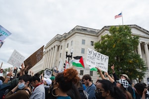 A group of protestors gather in front of a large government building holding various signs and flags. Signs contain messages supporting Palestine and opposing funding for genocide. The crowd is diverse, with individuals holding different types of protest signs, some featuring an American flag waving in the background.