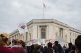 A group of people gather in front of a large, classical-style building, some holding up protest signs. The American flag flies from the top of the building. The mood is active with people engaged in a protest or demonstration.