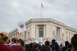 A group of people gather in front of a large, classical-style building, some holding up protest signs. The American flag flies from the top of the building. The mood is active with people engaged in a protest or demonstration.