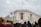 A group of people gather in front of a large, classical-style building, some holding up protest signs. The American flag flies from the top of the building. The mood is active with people engaged in a protest or demonstration.