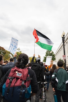 A group of people are participating in a protest. Several individuals are holding up signs; one reads 'CEASE FIRE' and another appears to have text related to ongoing issues. A large flag with red, black, white, and green colors is prominently displayed. The atmosphere is cloudy, and there are trees and buildings in the background.