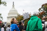 A crowd of people gather outside a government building, holding signs. The U.S. Capitol building is visible in the background. One prominent sign reads 'STOP WEAPONIZING OUR GRIEF.' The group appears to be part of a protest or rally, with individuals wearing varied clothing including a bright green jacket and a blue shirt.