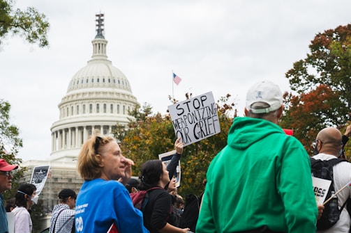 A crowd of people gather outside a government building, holding signs. The U.S. Capitol building is visible in the background. One prominent sign reads 'STOP WEAPONIZING OUR GRIEF.' The group appears to be part of a protest or rally, with individuals wearing varied clothing including a bright green jacket and a blue shirt.