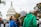 A crowd of people gather outside a government building, holding signs. The U.S. Capitol building is visible in the background. One prominent sign reads 'STOP WEAPONIZING OUR GRIEF.' The group appears to be part of a protest or rally, with individuals wearing varied clothing including a bright green jacket and a blue shirt.
