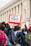 People are participating in a protest with a visible sign that reads 'CEASEFIRE NOW!' The crowd is dressed in casual clothing with backpacks visible, gathered in front of a classical-style building with columns.