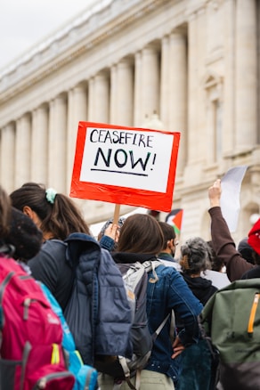 People are participating in a protest with a visible sign that reads 'CEASEFIRE NOW!' The crowd is dressed in casual clothing with backpacks visible, gathered in front of a classical-style building with columns.
