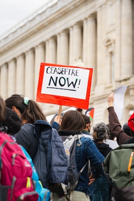 People are participating in a protest with a visible sign that reads 'CEASEFIRE NOW!' The crowd is dressed in casual clothing with backpacks visible, gathered in front of a classical-style building with columns.