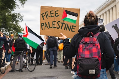A group of people gathered for a protest, with one person prominently holding a cardboard sign that reads 'Free Palestine'. The Palestinian flag is visible on the sign and other flags are present in the background. The crowd includes individuals with backpacks and bicycles, standing near a building with columns.