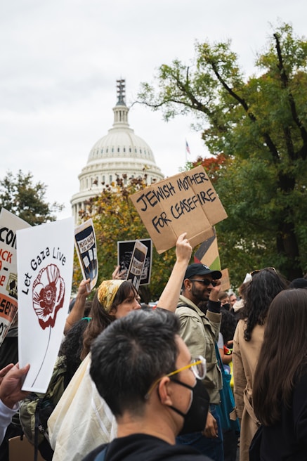 A crowd of people holding protest signs in front of a prominent white-domed building, with trees in the background. The signs display messages advocating for peace and justice.