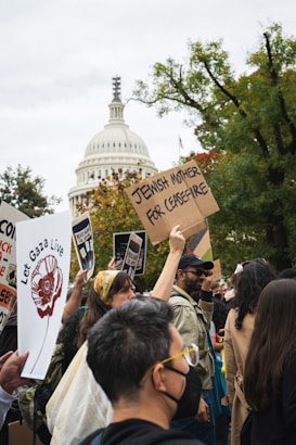 A crowd of people holding protest signs in front of a prominent white-domed building, with trees in the background. The signs display messages advocating for peace and justice.