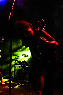 A musician is energetically playing an electric guitar on stage, with movements suggesting dynamic performance. The background features bright yellow and green stage lights and a drum set, enhancing the concert atmosphere. The scene is dark with spotlights creating vivid contrasts.