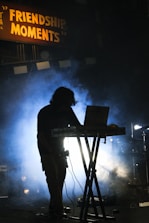 A person is silhouetted against bright stage lights while standing behind a music mixing console. Fog or smoke creates a dramatic effect, and a sign above reads 'FRIENDSHIP MOMENTS.' The setting suggests a musical performance or DJ event.