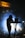 A person is silhouetted against bright stage lights while standing behind a music mixing console. Fog or smoke creates a dramatic effect, and a sign above reads 'FRIENDSHIP MOMENTS.' The setting suggests a musical performance or DJ event.