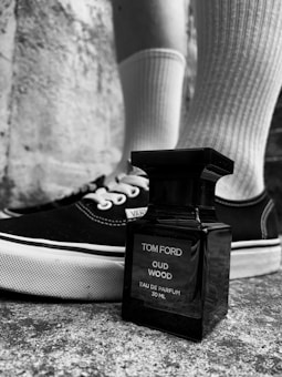 A black and white photograph featuring a Tom Ford Oud Wood perfume bottle positioned on a textured stone surface. Behind the bottle, a pair of black Vans sneakers with white soles is visible, along with legs wearing white ribbed socks. The overall composition is stylish and moody.
