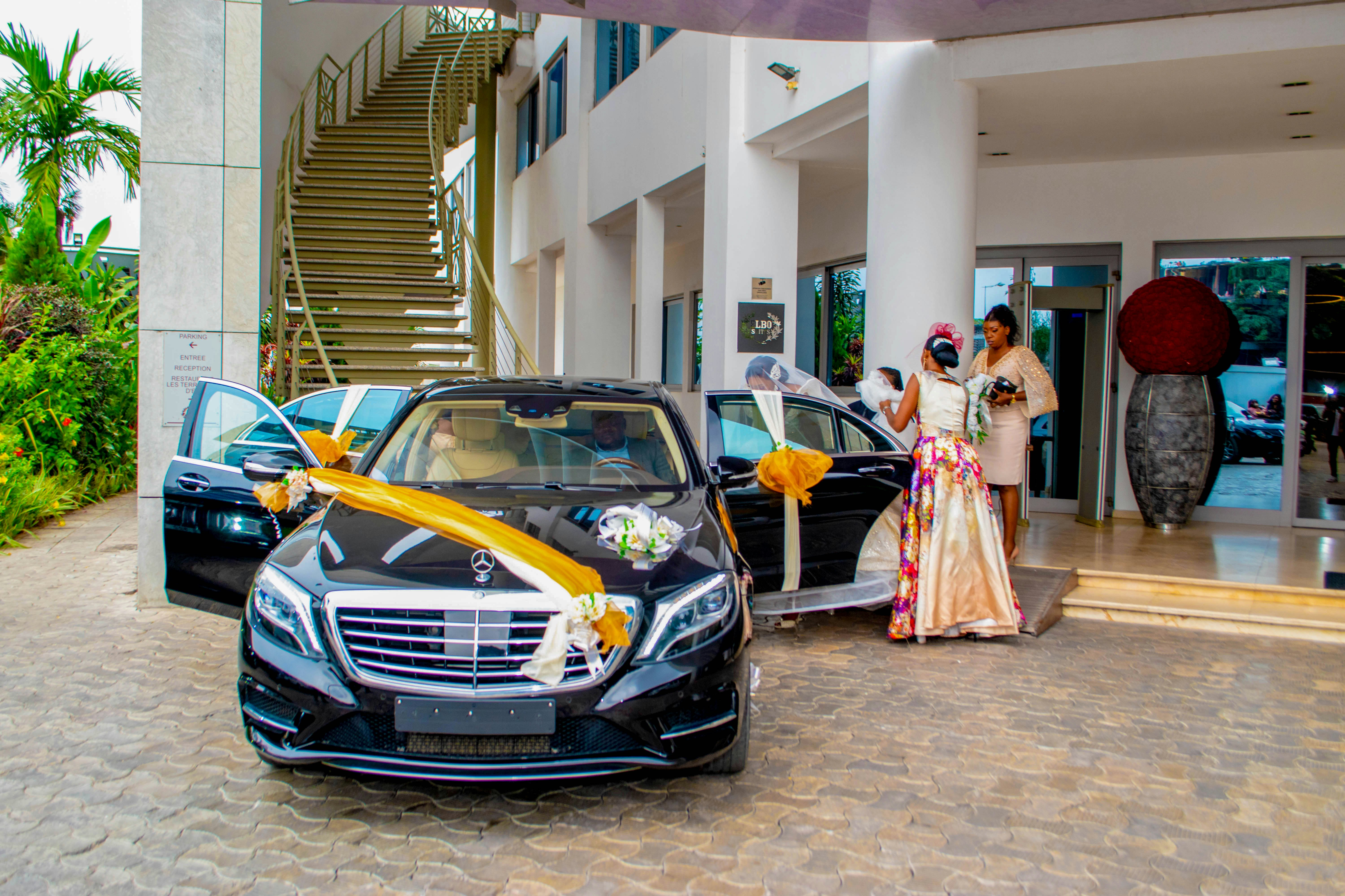 Elegant black luxury sedan decorated with white ribbons and flowers parked at upscale wedding venue with romantic garden setting and string lights