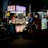 A dimly lit street food stall with vibrant signage offers a selection of snacks. A person wearing a helmet is seated on a scooter, waiting in front of the stall, while another person is busy inside preparing food. The scene is lively with bright lights and colorful stalls creating a night market ambiance.