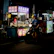 A dimly lit street food stall with vibrant signage offers a selection of snacks. A person wearing a helmet is seated on a scooter, waiting in front of the stall, while another person is busy inside preparing food. The scene is lively with bright lights and colorful stalls creating a night market ambiance.