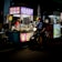 A dimly lit street food stall with vibrant signage offers a selection of snacks. A person wearing a helmet is seated on a scooter, waiting in front of the stall, while another person is busy inside preparing food. The scene is lively with bright lights and colorful stalls creating a night market ambiance.