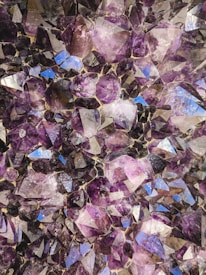 A close-up view of a cluster of amethyst crystals, displaying various shades of purple with some reflective surfaces catching light and creating a shimmering effect. The crystals have angular facets and are tightly packed together, revealing the intricate structure of the mineral formation.