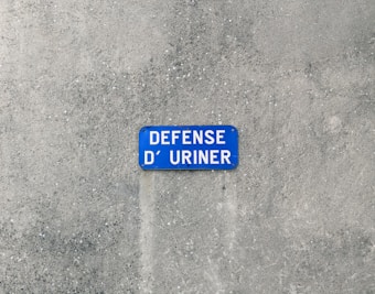 A blue sign with white lettering is mounted on a textured gray wall. The sign contains the French words 'DÉFENSE D’ URINER,' indicating a prohibition against urination.