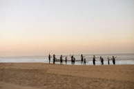 Group of travelers practicing guided breathing exercises by the sea at dawn.