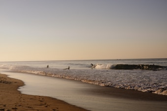A tranquil beach scene with gentle waves rolling onto a sandy shore. Several surfers are in the water, with one riding a wave. The sky is clear, and the lighting suggests either early morning or late afternoon, casting a warm glow.