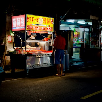 A street food vendor is preparing food at a brightly lit stall. One customer stands in front of the cart, and the vendor is wearing a mask while handling the food. The street is dimly lit, and there is a sense of nighttime ambiance.
