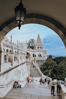 A scenic view captures a historical stone architecture with arches and a conical tower surrounded by trees. People are walking and sitting on the steps, enjoying the scenery. The sky is partly cloudy, and a lantern hangs prominently in the foreground.