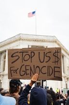 A crowd holds a large cardboard sign with the message 'Stop US $ For Genocide' in front of a building with a U.S. flag on top. The scene suggests a protest or demonstration, with people gathered closely and focused on the action.