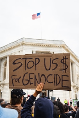 A crowd holds a large cardboard sign with the message 'Stop US $ For Genocide' in front of a building with a U.S. flag on top. The scene suggests a protest or demonstration, with people gathered closely and focused on the action.