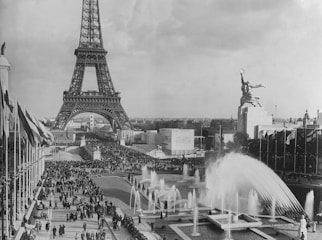 A historical black and white photograph of the Eiffel Tower surrounded by a large crowd of people. Fountains with water spraying are prominent in the foreground, and flags line both sides of a walkway.