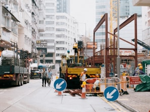 A busy urban construction site with workers in helmets performing tasks. Various machinery, including a crane, is visible, and several vehicles are parked nearby. The surrounding area consists of tall buildings, and traffic is present, contributing to the vibrant city atmosphere. Temporary barriers and signs indicate ongoing work, and a footpath is closed.