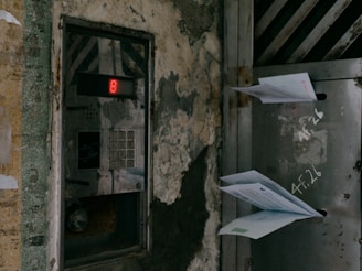 An old, weathered intercom system is mounted on a wall with visible decay and peeling paint. A digital display shows the number 8, and beneath it is a keypad. Adjacent to the intercom, a metal mailbox is attached to the wall, with several letters protruding from the slot. Graffiti is scrawled on the wall, displaying some illegible words or numbers.