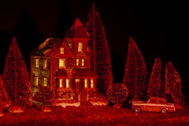 A miniature scene featuring a detailed model of a two-story house with a steep roof, illuminated from within. It is surrounded by evenly spaced, conically shaped evergreen trees, covered in a dusting of white that resembles snow. In front of the house, a small vintage car model is parked. The entire scene is bathed in a warm, deep red light, creating a dramatic and cozy ambiance.