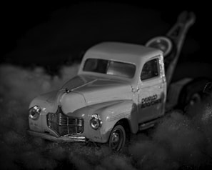 A vintage toy tow truck is placed on a fluffy surface, with intricate details visible on the model, such as the front grille and headlights. The image is in black and white, lending a nostalgic and classic feel.