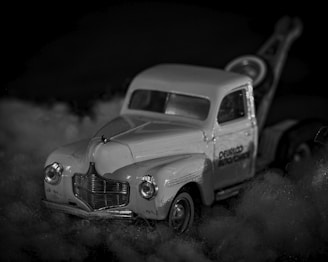 A vintage toy tow truck is placed on a fluffy surface, with intricate details visible on the model, such as the front grille and headlights. The image is in black and white, lending a nostalgic and classic feel.