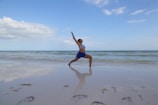 A person is striking a yoga pose on a sandy beach with gentle ocean waves in the background. The sky is mostly clear with a few clouds, and the reflection of the person is visible on the wet sand.