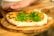 A freshly baked pizza topped with arugula placed on a wooden board. The crust appears golden with some charred spots, and there is a hand visible in the background.