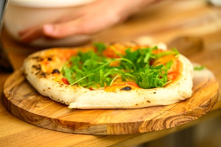 A freshly baked pizza topped with arugula placed on a wooden board. The crust appears golden with some charred spots, and there is a hand visible in the background.