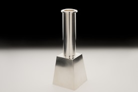 A metallic candlestick with a sleek, modern design featuring a cylindrical shaft and a square base. It is positioned against a black to white gradient background, highlighting its reflective surface.