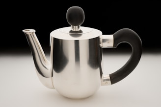 A sleek, modern stainless steel teapot with a glossy finish sits on a smooth surface. It features a black handle and knob, creating a striking contrast with its metallic body. The spout and lid are seamlessly integrated into the design.