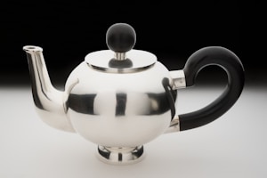 A shiny metal teapot with a modern, rounded design features a reflective silver surface. It has a sleek spout and a unique black handle shaped in an elegant curve. The lid is adorned with a black spherical knob.