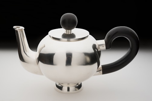 A shiny metal teapot with a modern, rounded design features a reflective silver surface. It has a sleek spout and a unique black handle shaped in an elegant curve. The lid is adorned with a black spherical knob.