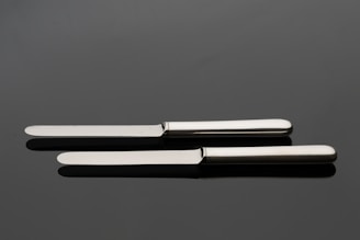 A pair of silver butter knives positioned parallel on a reflective dark surface.